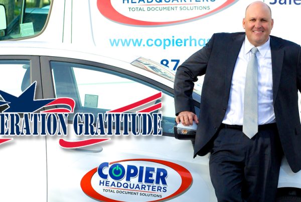 A man in a suit smiles while leaning on a car with a Copier Headquarters logo, representing the Los Angeles firm. Behind him is a branded van. The text Operation Gratitude with a star and flag graphic is overlaid on the image.