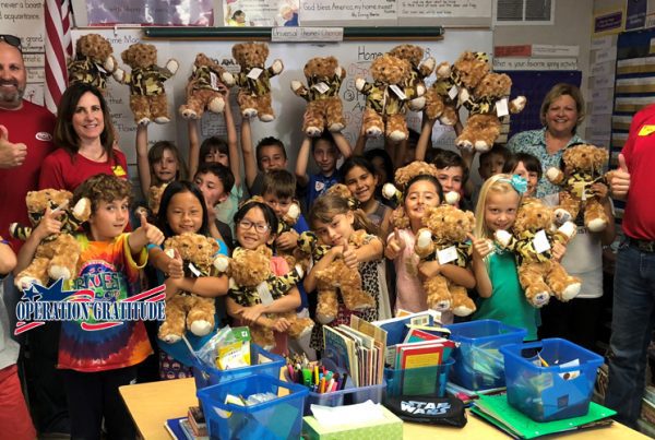 A group of smiling children and adults in a colorful classroom hold up teddy bears. Artwork decorates the walls, books fill the desks, and an Operation Gratitude banner is proudly displayed in the foreground.