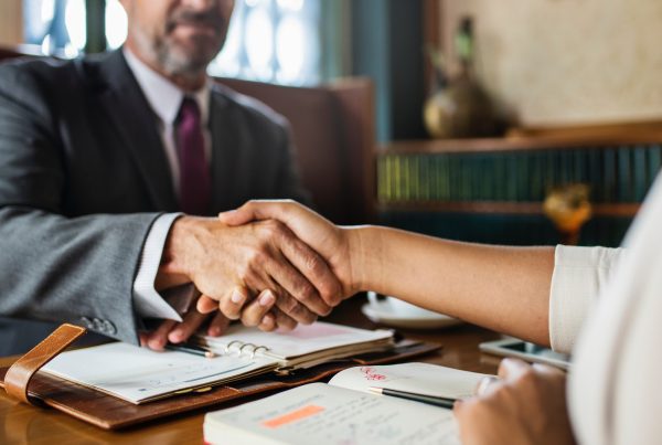 Two people are shaking hands across a desk with open notebooks and paperwork, suggesting a business meeting or agreement—perhaps an acquisition discussion involving Advanced Office Automation—in a professional GoodSuite office environment.