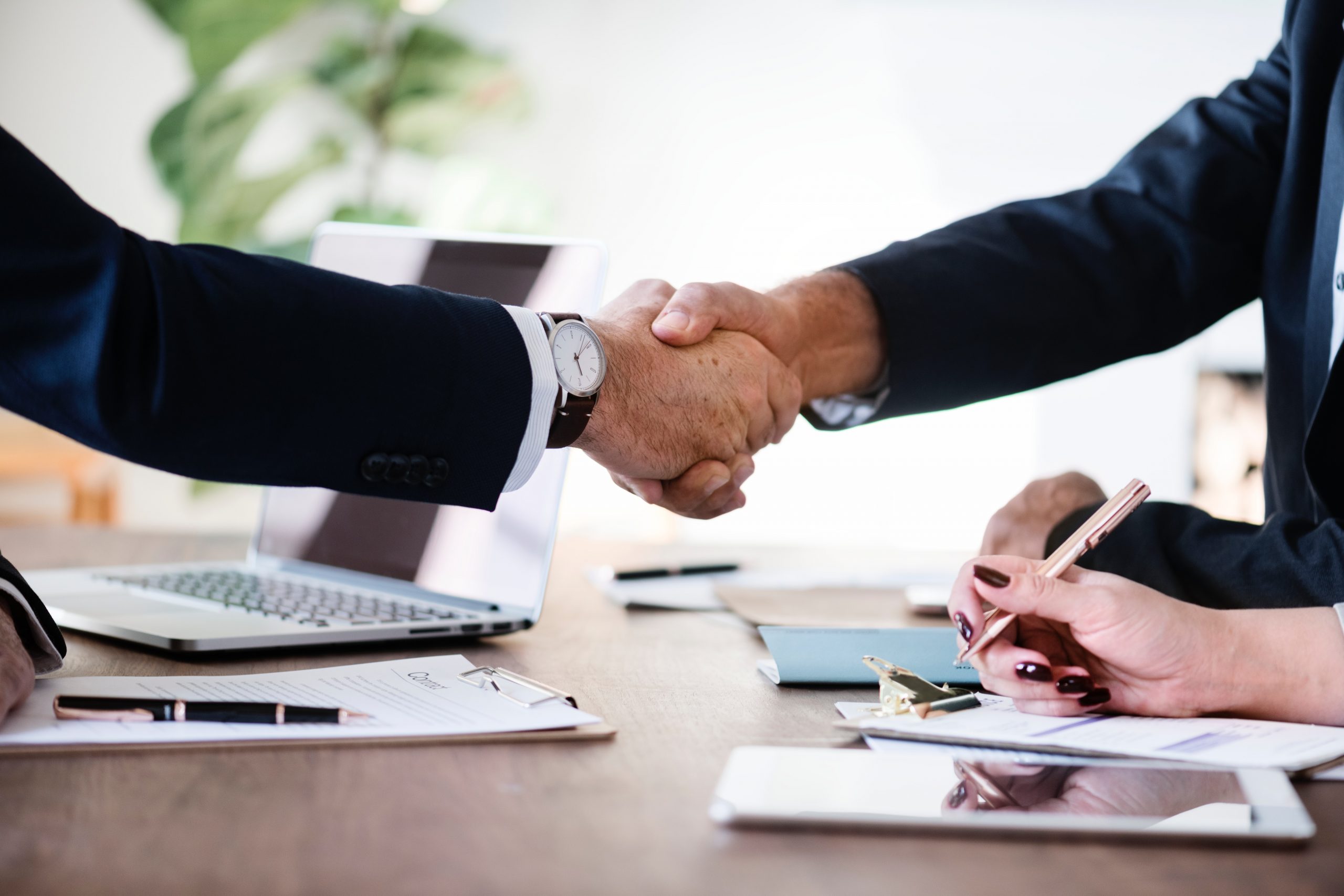 Two people in business attire shake hands across a desk—documents, a laptop, and a tablet rest nearby—as a third person from Office Machine Specialists takes notes during an office machine acquisition meeting.