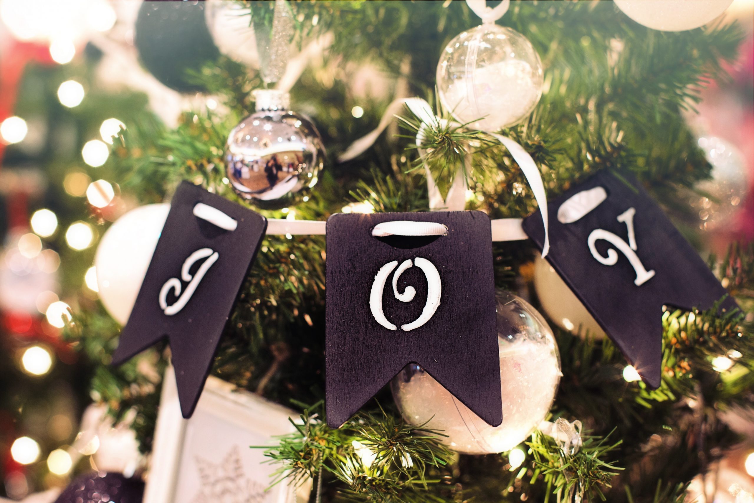 A close-up of a decorated Christmas tree with black banners spelling JOY in white letters, surrounded by ornaments and glowing lights, captures the spirit of the holidays and giving back.