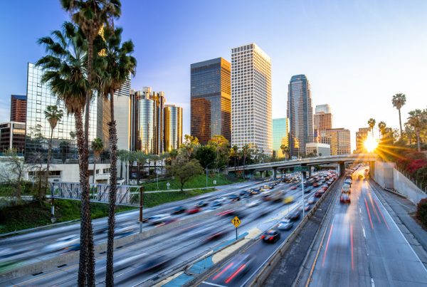 A busy freeway with blurred cars at sunset runs through downtown Los Angeles, lined with tall palm trees and modern skyscrapers reflecting the golden light—home to thriving businesses and copy machine leasing services.