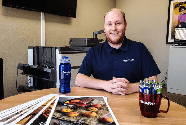 A smiling Summer Intern in a navy polo shirt sits at a round office table with printed photos, a blue water bottle, and a mug filled with pens. Office equipment and a framed poster are in the background.
