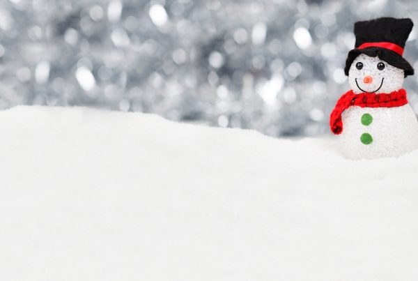 A small snowman with a black top hat, red scarf, green buttons, and a smiling face stands on a snowy surface with a blurred, sparkling silver background, capturing the festive spirit of Holiday Drive 2019.