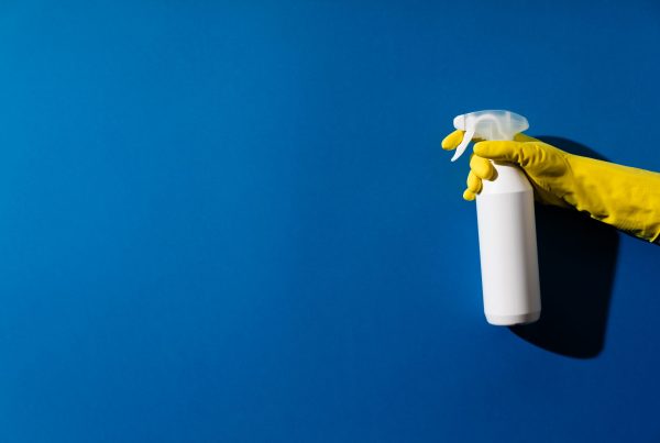 A hand wearing a yellow rubber glove holds a white spray bottle against a solid blue background, casting a shadow to the right—a nod to GoodSuite's commitment to office response and safety in the wake of COVID-19.