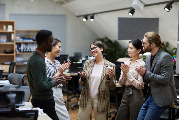 Five colleagues stand in an office, smiling and clapping, as they celebrate a Promotion Announcement in a friendly and professional atmosphere.
