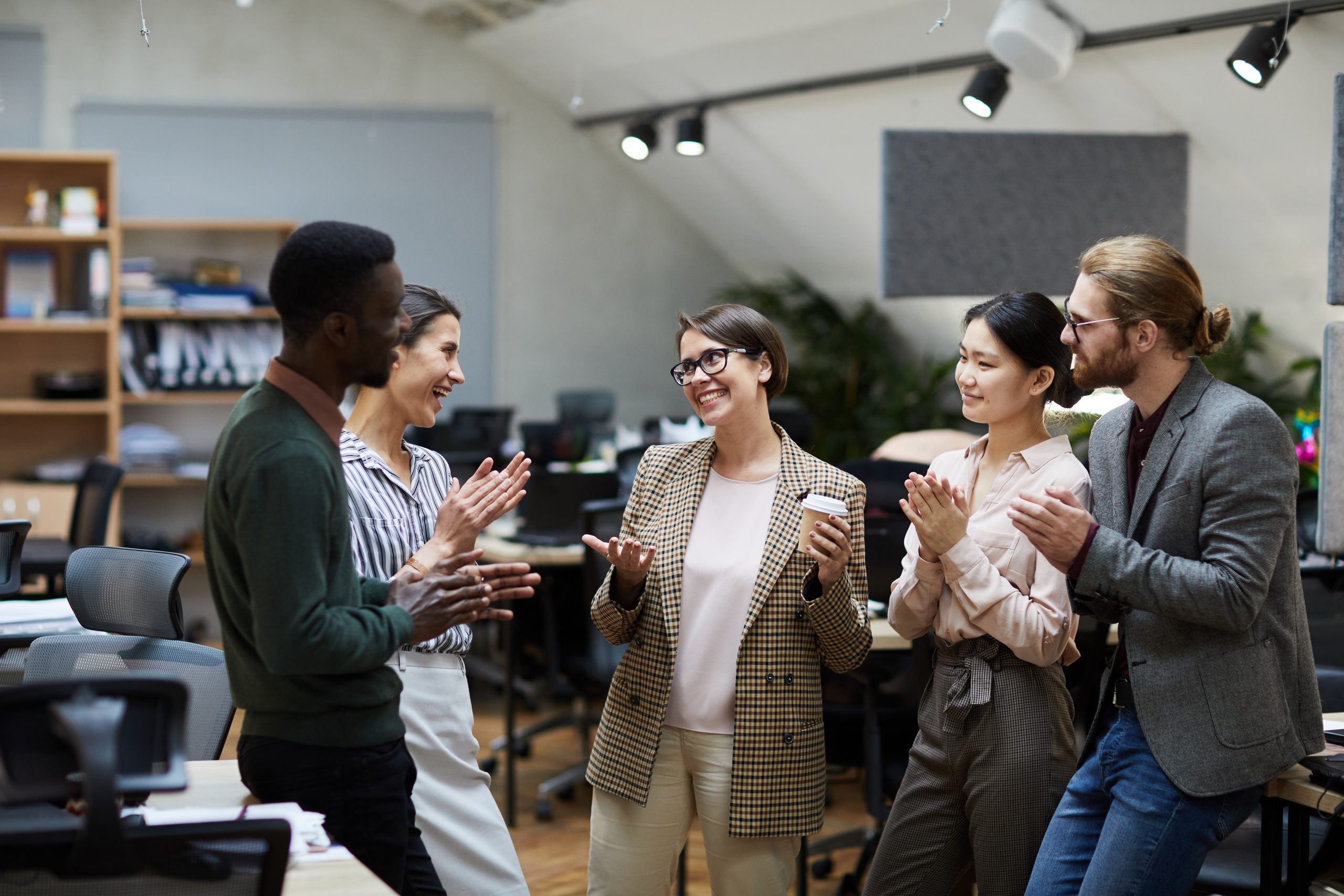 Five colleagues stand in an office, smiling and clapping, as they celebrate a Promotion Announcement in a friendly and professional atmosphere.