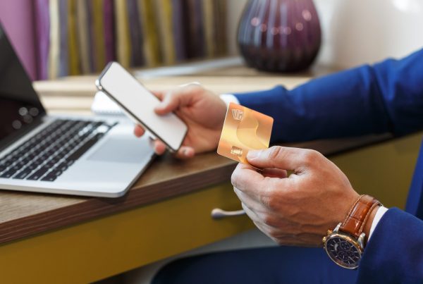 A person in a blue suit holds a smartphone and a gold credit card while sitting at a desk with a laptop, preparing to make a one-time payment online. A lamp and files are visible in the background.