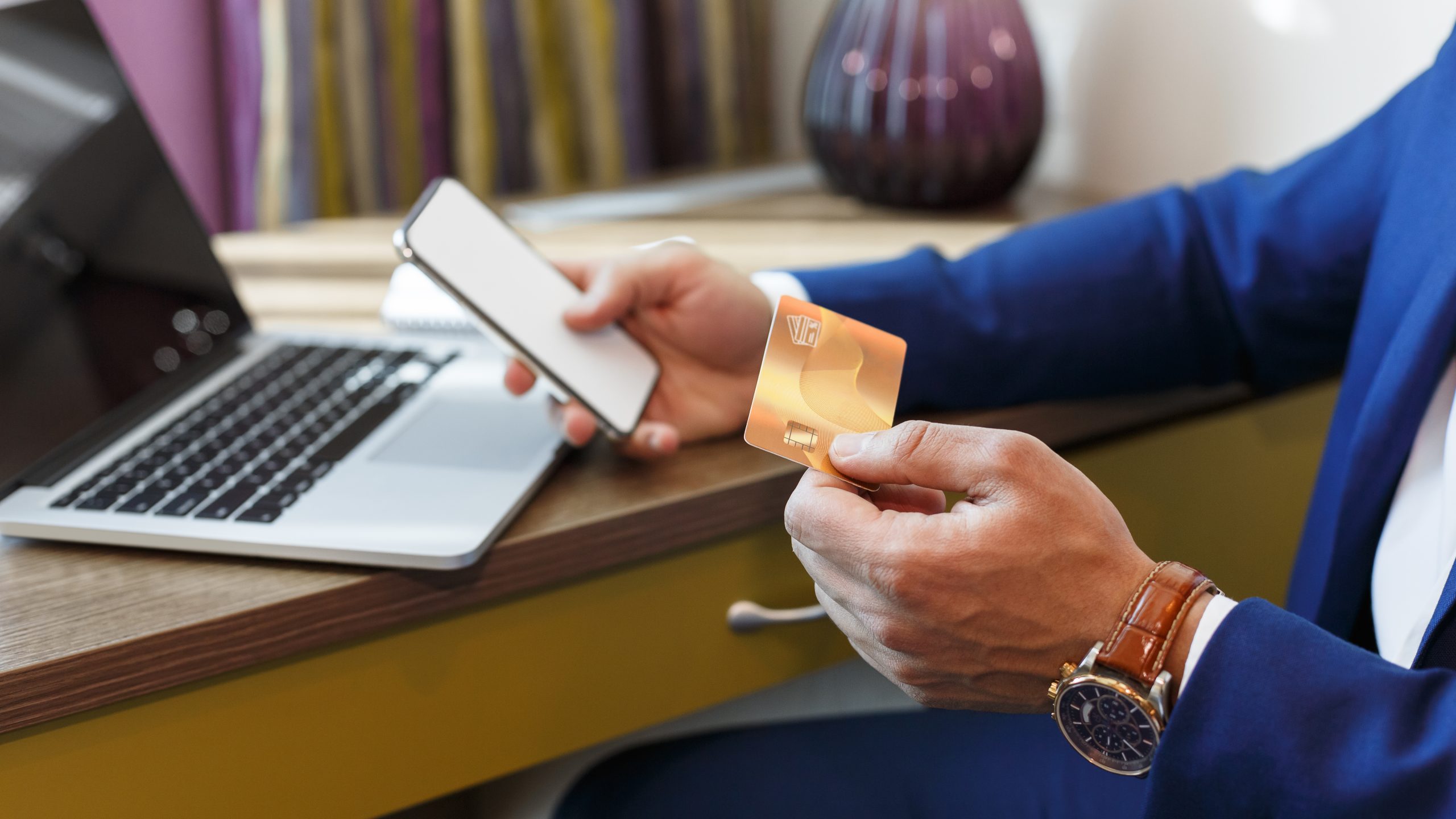 A person in a blue suit holds a smartphone and a gold credit card while sitting at a desk with a laptop, preparing to make a one-time payment online. A lamp and files are visible in the background.
