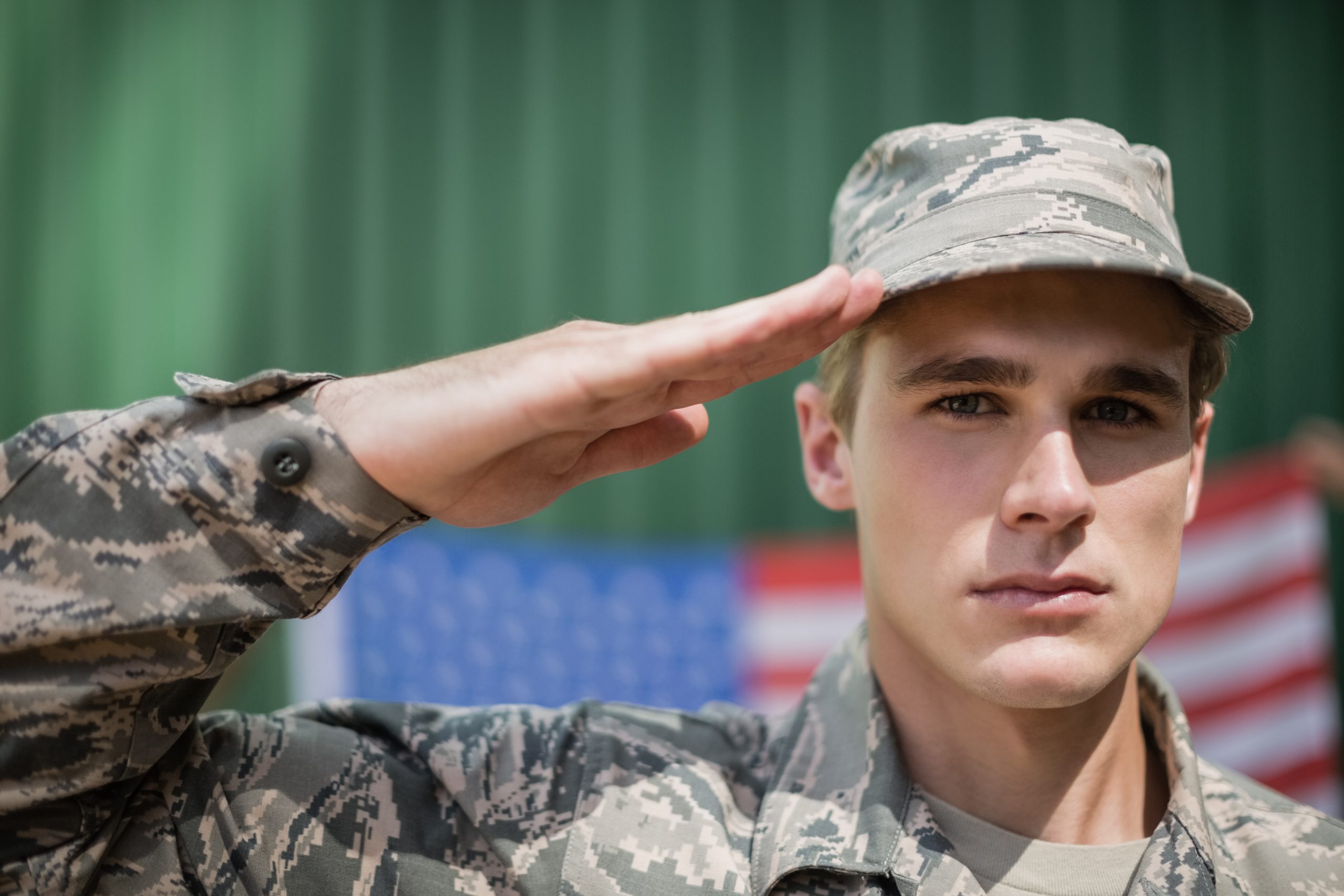 A young man in military uniform salutes while standing in front of a blurred American flag backdrop, honoring Military Appreciation Day.