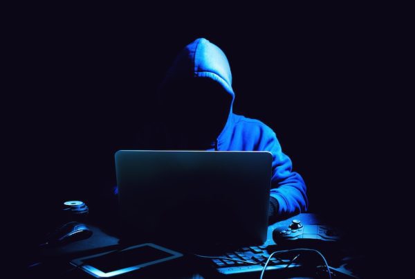 A person in a blue hoodie, face obscured by shadows, sits in a dark room like many hackers, using a laptop, with a tablet, mouse, game controller, and keyboard scattered on the desk before them.
