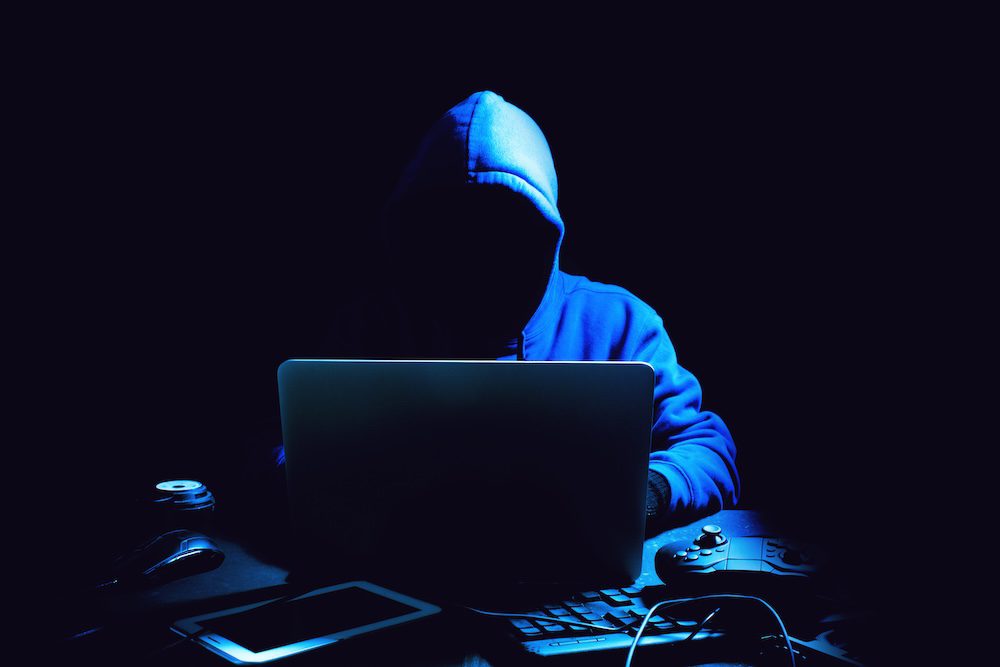 A person in a blue hoodie, face obscured by shadows, sits in a dark room like many hackers, using a laptop, with a tablet, mouse, game controller, and keyboard scattered on the desk before them.