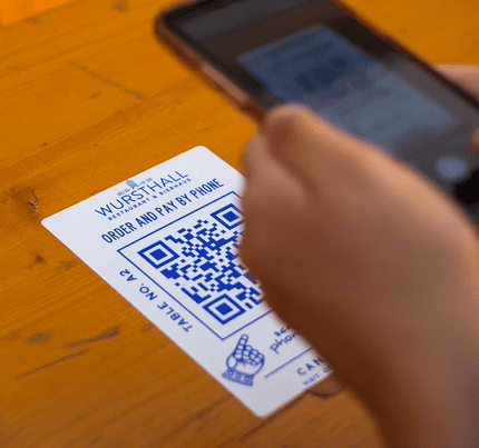 A person demonstrates the effective use of QR codes by holding a smartphone over a code printed on a WURSTHALL card to order and pay, showcasing how print materials can streamline dining at a wooden table.