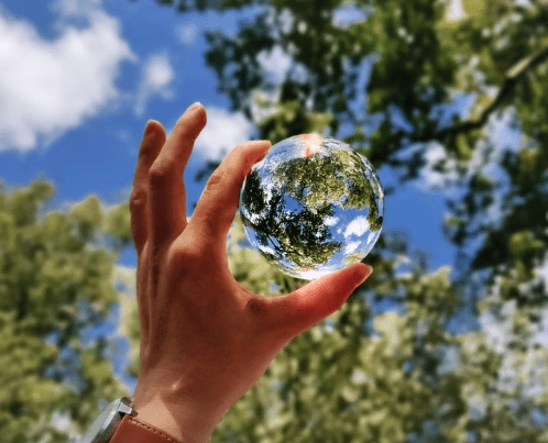 A hand wearing a brown watch holds up a clear glass sphere that reflects trees and the blue sky, emphasizing the beauty of the environment against a blurred background of leafy green trees.