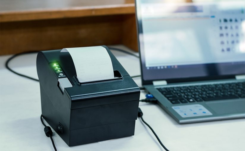 A black thermal receipt printer with a printed receipt sits on a desk next to an open laptop. Both devices are plugged in and connected, showcasing a great choice for anyone consulting a printer buying guide.