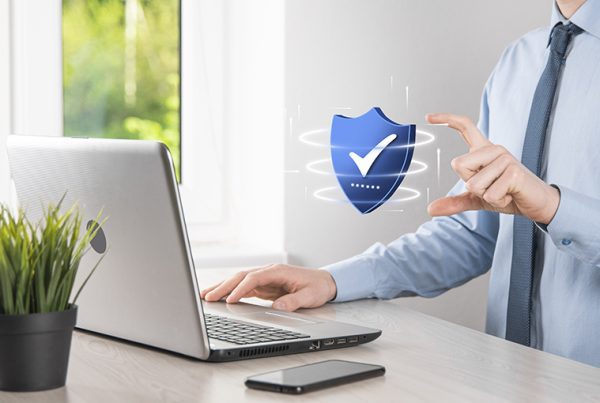 A person in business attire sits at a desk with a laptop, touching a floating digital shield with a check mark, symbolizing essential cybersecurity facts. A potted plant and smartphone are also on the desk.