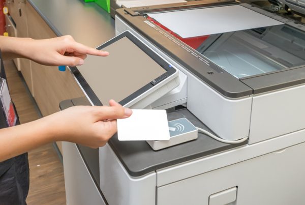 A person uses a white access card and touches a scanner on a photocopier, while the other hand operates the touchscreen panel—demonstrating steps from a print security checklist to enhance print security.