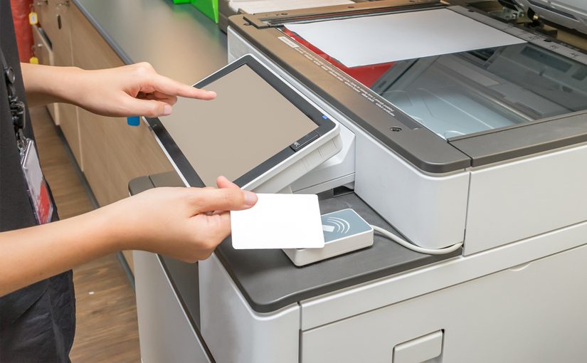 A person uses a white access card and touches a scanner on a photocopier, while the other hand operates the touchscreen panel—demonstrating steps from a print security checklist to enhance print security.