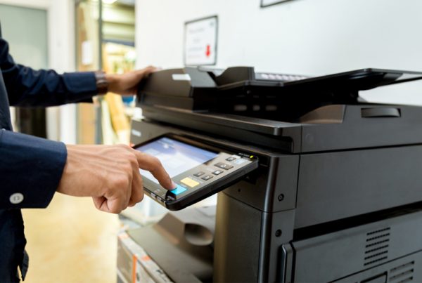 A person in a dark shirt uses the touchscreen panel on a black printer in an office setting, following a Basic Guide for Managed Print Services.