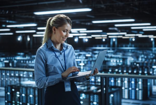 A woman in business attire stands in a modern data center, smiling while using a laptop. Rows of illuminated server racks fill the large, high-tech room—highlighting the efficiency of Managed IT Services in 2022.