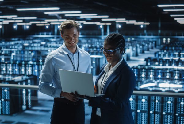 Two professionals stand in a large, modern server room, smiling and looking at a laptop as they conduct a cybersecurity assessment. Rows of illuminated server racks fill the background, creating a high-tech atmosphere.