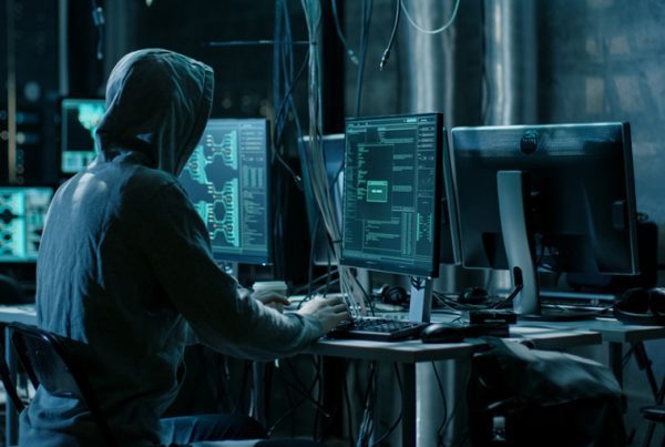 A person wearing a hoodie sits at a desk with multiple computer monitors displaying code and technical data in a dimly lit room, highlighting the importance of Dark Web Security in today’s cybersecurity or hacking environment.