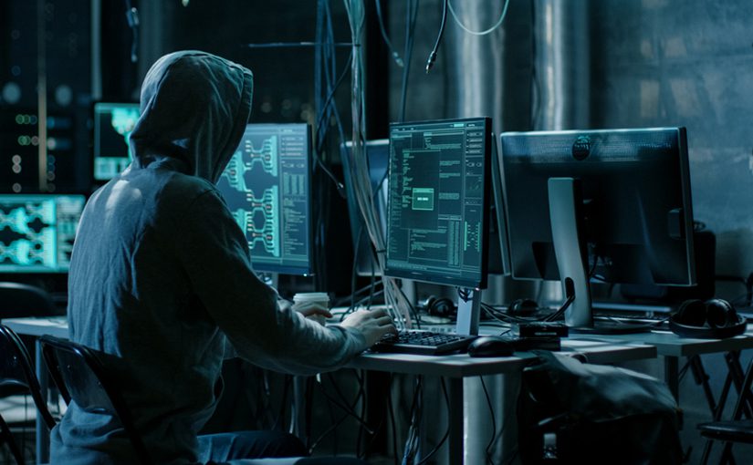 A person wearing a hoodie sits at a desk with multiple computer monitors displaying code and technical data in a dimly lit room, highlighting the importance of Dark Web Security in today’s cybersecurity or hacking environment.