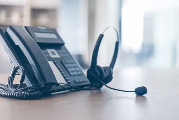 A black office desk phone with a corded handset sits on a wooden desk next to a black headset with a microphone, showcasing modern VoIP systems in a bright office setting.