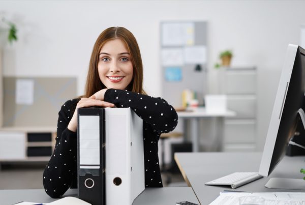 A woman with long brown hair, wearing a black polka dot sweater, sits at a desk in an office, smiling and resting her arms on two large binders—showcasing her expert office organization tips for 2023—with a computer monitor beside her.
