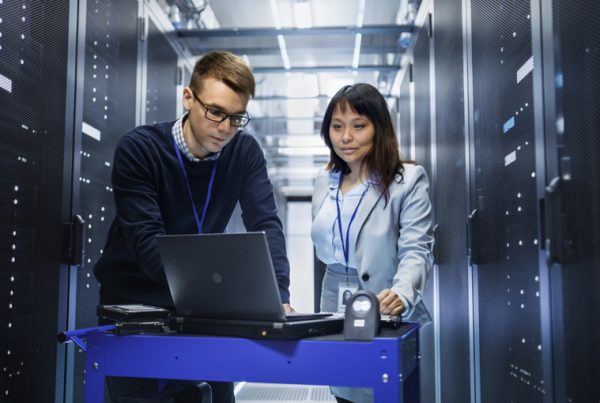 Two IT professionals, a man and a woman, stand in a server room looking at a laptop on a cart, delivering Managed IT Services. They wear ID badges and are surrounded by tall server racks with visible cables and lights.