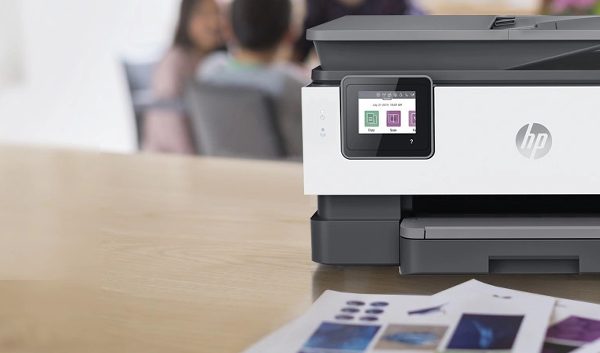 An HP printer, featured among the Best Printers 2023, sits on a wooden desk with colorful documents in front. In the blurred background, people are seated at a table, suggesting an office or collaborative workspace.