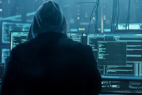 A person wearing a hoodie sits in front of multiple computer monitors displaying code, digital maps, and data in a dark room with a blue tint, suggesting a Dark Web Monitoring or cybersecurity environment focused on business protection.