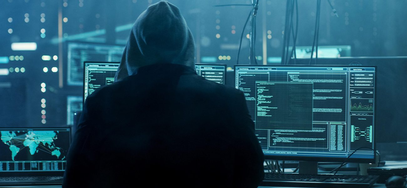 A person wearing a hoodie sits in front of multiple computer monitors displaying code, digital maps, and data in a dark room with a blue tint, suggesting a Dark Web Monitoring or cybersecurity environment focused on business protection.