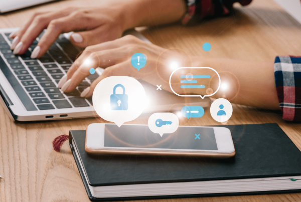 A person types on a laptop at a wooden desk with a smartphone on a notebook nearby. Digital icons appear above the phone, representing cybersecurity tips for small business owners.