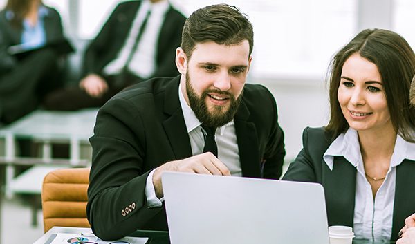 Three business professionals in suits sit together at a desk, looking at a laptop screen and discussing how to manage business IT needs. Other people are seated and talking in the background in a modern office setting.