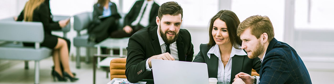 Three business professionals in suits sit together at a desk, looking at a laptop screen and discussing how to manage business IT needs. Other people are seated and talking in the background in a modern office setting.
