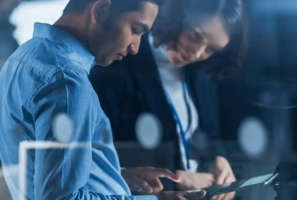 Two people in business attire are standing close together in a modern office, discussing information on a digital tablet—highlighting collaboration and decision-making around managed IT services. Blurred lights and office equipment form the backdrop.