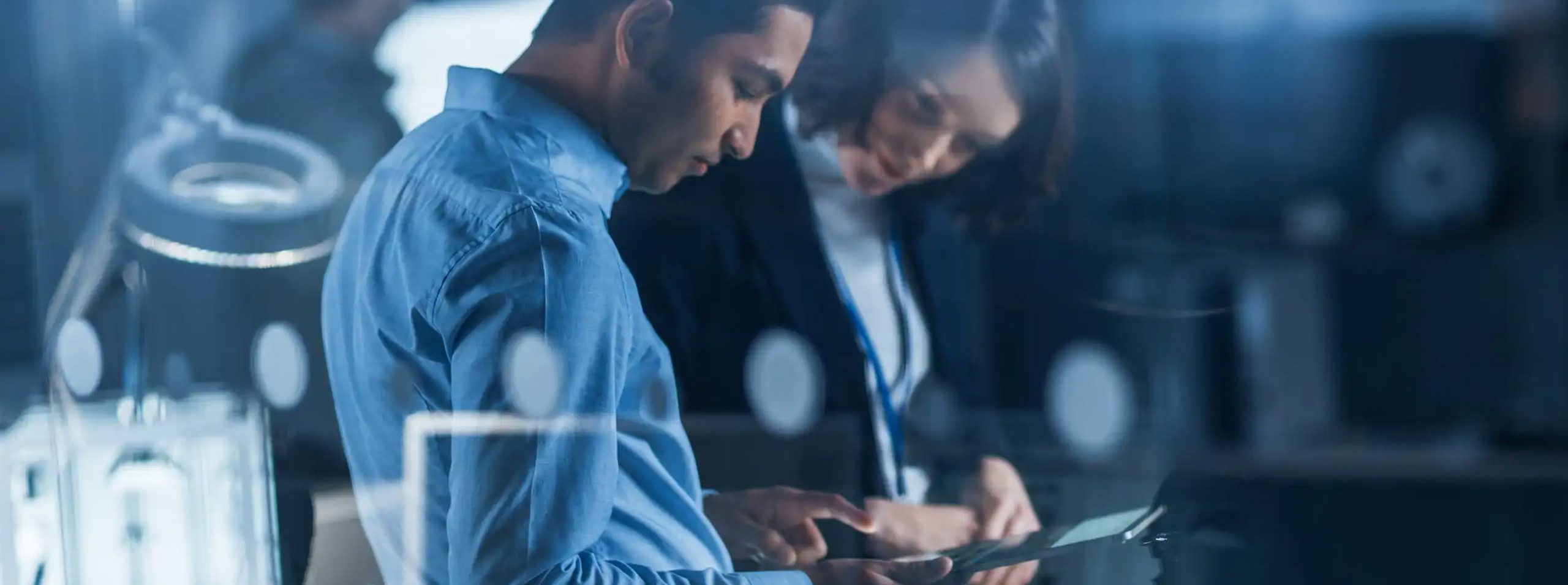 Two people in business attire are standing close together in a modern office, discussing information on a digital tablet—highlighting collaboration and decision-making around managed IT services. Blurred lights and office equipment form the backdrop.