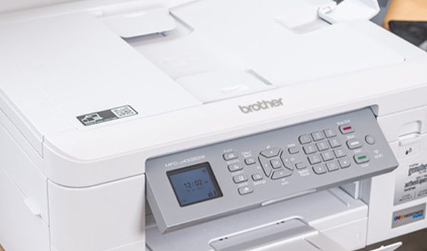 A white Brother printer sits on a wooden desk next to another printer, with a person at a workstation—ideal for offices using Managed Print Services to streamline print management alongside their everyday tech.