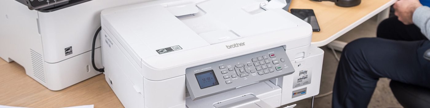 A white Brother printer sits on a wooden desk next to another printer, with a person at a workstation—ideal for offices using Managed Print Services to streamline print management alongside their everyday tech.