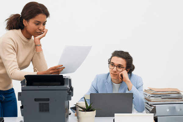 Three women in an office, one standing and examining a paper, while the other two sit at a cluttered desk with a laptop, looking thoughtful and focused—collaborating on print services for small businesses among office equipment and paperwork.