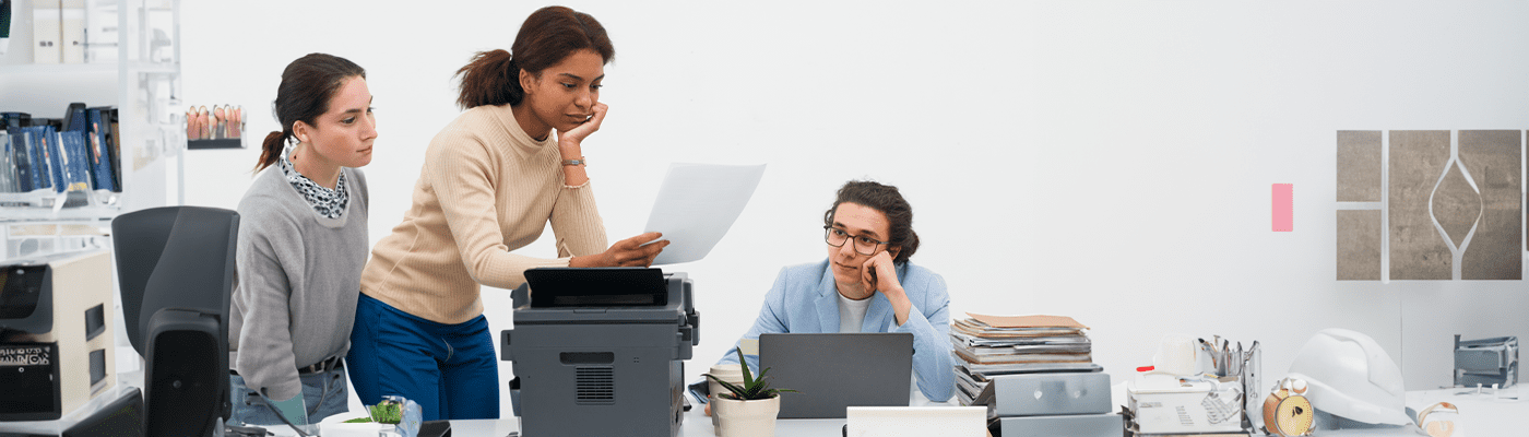 Three women in an office, one standing and examining a paper, while the other two sit at a cluttered desk with a laptop, looking thoughtful and focused—collaborating on print services for small businesses among office equipment and paperwork.