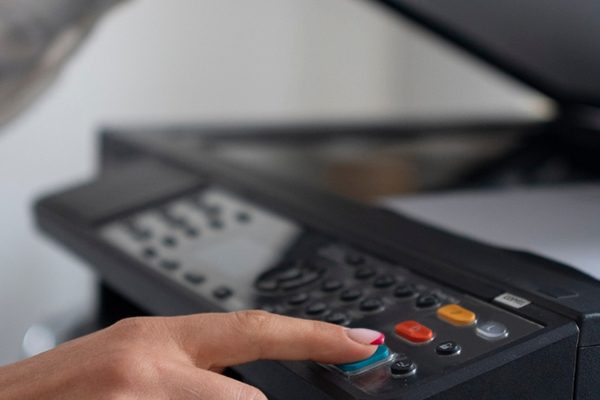 A person presses a button on a photocopier or printer, with papers placed on the scanner bed and the lid open, demonstrating efficient office printing.