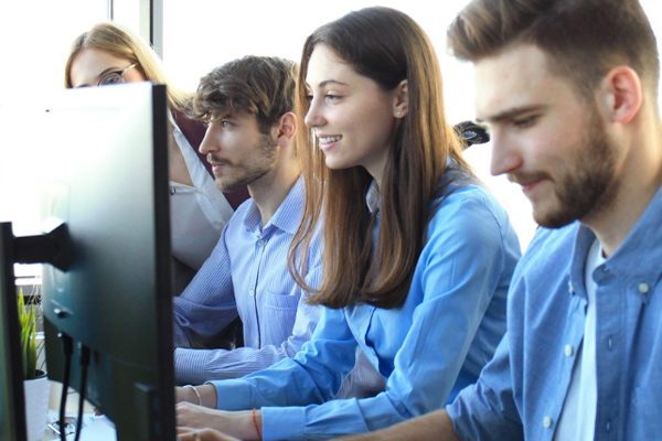 Four young adults sit in a row at desks, working on computers in a bright, modern business office. One person stands behind them, appearing to offer guidance about print services and when to get assistance.