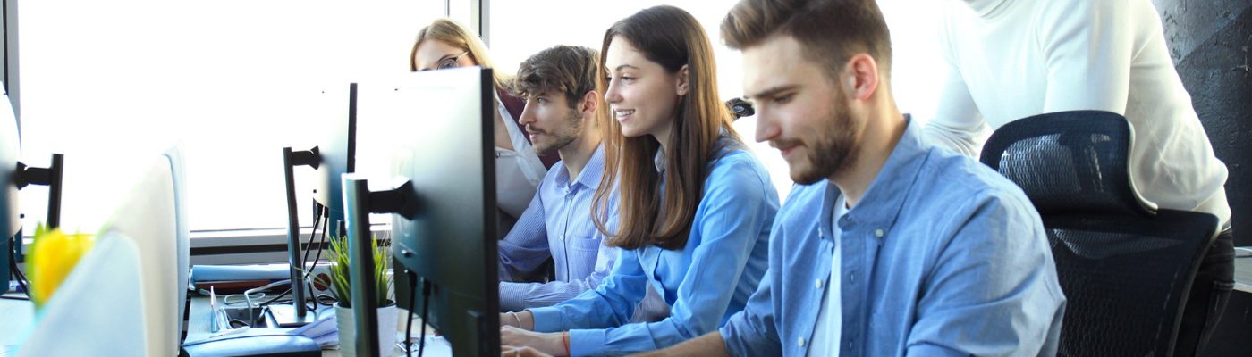 Four young adults sit in a row at desks, working on computers in a bright, modern business office. One person stands behind them, appearing to offer guidance about print services and when to get assistance.