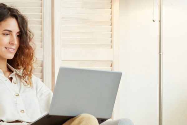 A woman with curly hair sits in a cozy chair, using a laptop and wearing wireless earbuds. She appears to be smiling, with bookshelves and a desk in the background, embodying the empowered remote workforce in a comfortable home office setting.