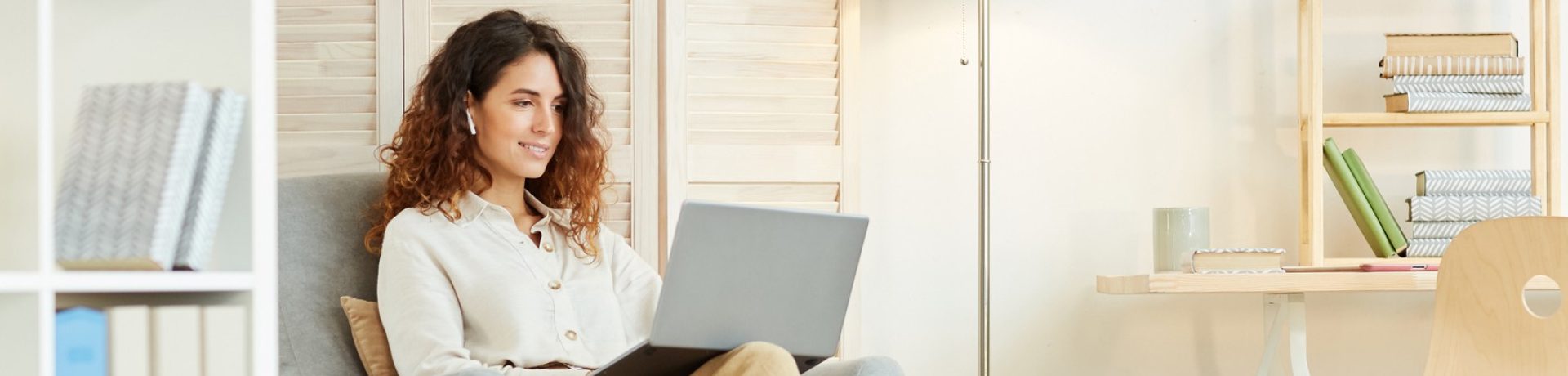 A woman with curly hair sits in a cozy chair, using a laptop and wearing wireless earbuds. She appears to be smiling, with bookshelves and a desk in the background, embodying the empowered remote workforce in a comfortable home office setting.