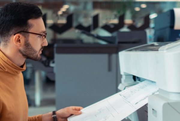 A person wearing glasses and a brown turtleneck retrieves a large print and copy document from a wide-format printer in a modern office environment. Shelves with paper rolls are visible in the background, supporting evolving business needs.