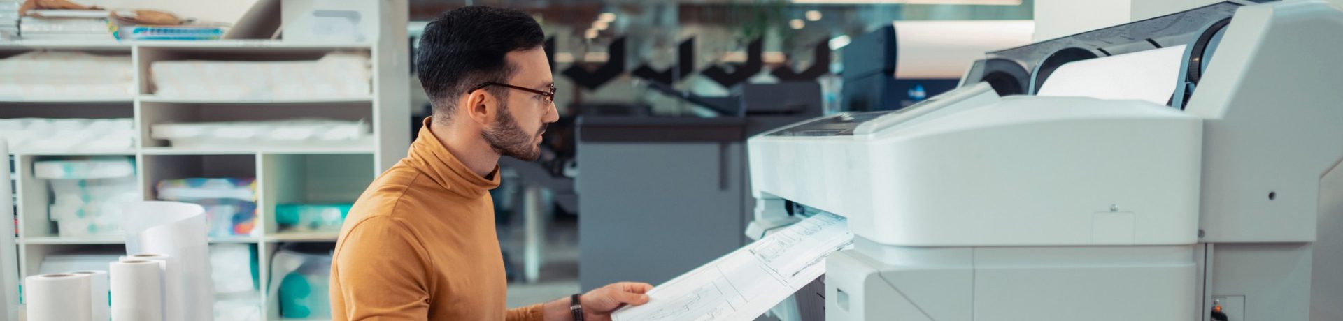A person wearing glasses and a brown turtleneck retrieves a large print and copy document from a wide-format printer in a modern office environment. Shelves with paper rolls are visible in the background, supporting evolving business needs.