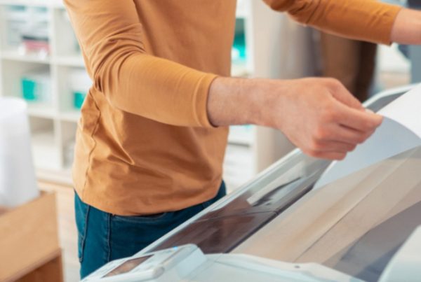 A person in a brown long-sleeve shirt operates a large format printer, feeding paper into the machine. Rolls of paper are visible in the background, highlighting business efficiency in a busy print services studio.